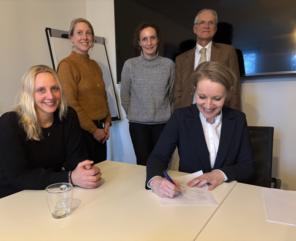 Foto van tekenmoment. 5 blije personen op de foto, één daarvan ondertekent het document, de rest kijkt mee. 